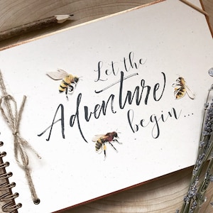 May include: A cream-coloured spiral-bound journal with the phrase "Let the Adventure begin..." in elegant script. The cover features bee illustrations. A pencil and twine are visible. The journal has a copper-coloured edge.