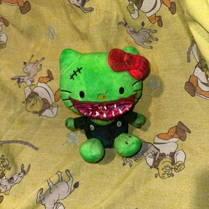 May include: Green plush toy with a red bow and a large, toothy grin. The toy is a Hello Kitty character with a stitched-up scar on its forehead.