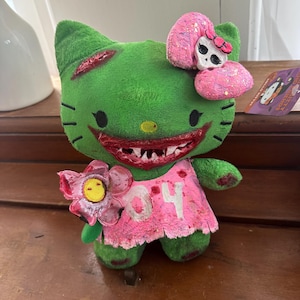 May include: Green plush toy wearing a pink dress and a pink bow with a skull on top. The toy has a large, toothy grin and a pink flower in its hand. The toy is labelled "04".