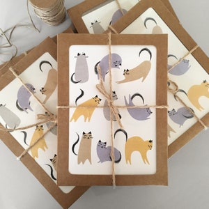 May include: Set of six greeting cards featuring illustrations of cats in various poses. The cards are printed on white cardstock and have a brown kraft paper envelope. The cards are tied together with twine.