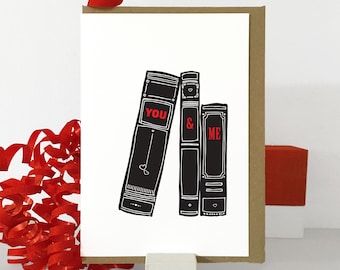 Può includere: Un biglietto bianco con illustrazioni in bianco e nero di tre libri. I libri hanno le parole "YOU" e "& ME" scritte in rosso.