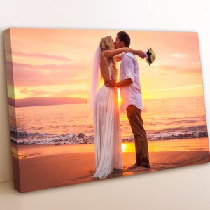 May include: A couple in wedding attire kiss on a beach at sunset. The bride is wearing a white gown and veil, and the groom is wearing a white shirt and khaki pants. The couple is standing on a sandy beach with the ocean in the background. The sky is a vibrant orange and pink.