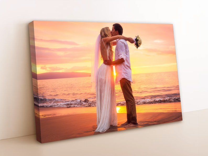 May include: A couple in wedding attire kiss on a beach at sunset. The bride is wearing a white gown and veil, and the groom is wearing a white shirt and khaki pants. The couple is standing on a sandy beach with the ocean in the background. The sky is a vibrant orange and pink.