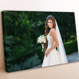 May include: A bride in a white wedding dress with a long veil, holding a bouquet of white flowers. She is standing in a garden with green trees and bushes in the background.