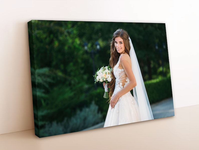 May include: A bride in a white wedding dress with a long veil, holding a bouquet of white flowers. She is standing in a garden with green trees and bushes in the background.
