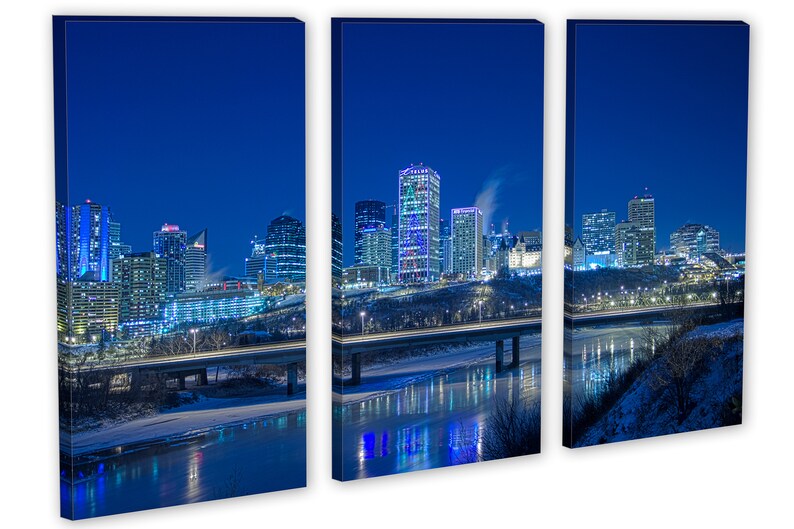 May include: A triptych canvas print of a city skyline at night. The buildings are illuminated with various colors, reflecting in the water below. A bridge with car lights crosses the river. The sky is a deep blue.