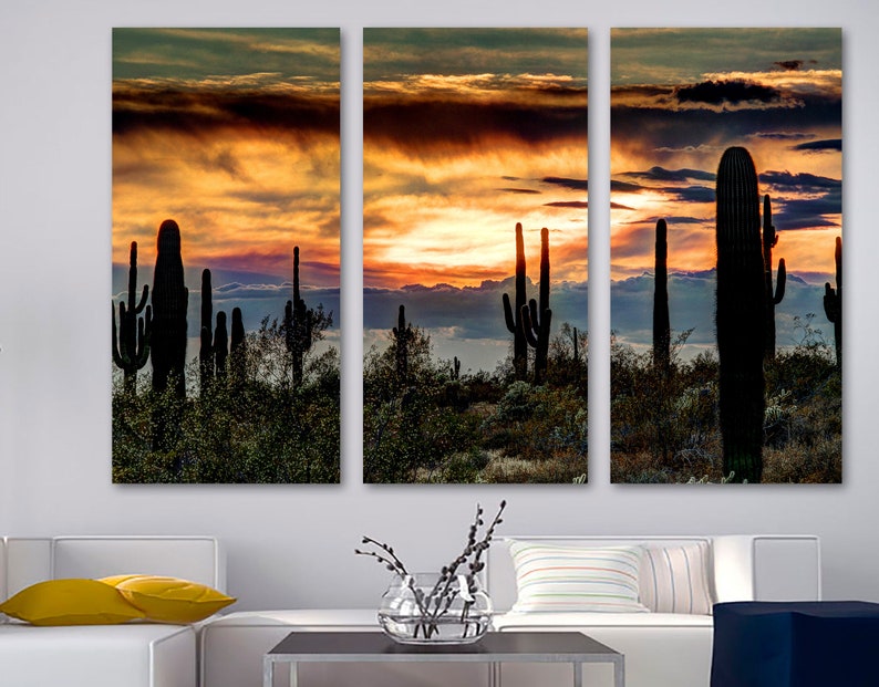 May include: A triptych canvas print featuring a desert landscape at sunset. Silhouetted saguaro cacti and other desert plants are set against a vibrant sky of orange, yellow, and dark blue hues. The artwork evokes a sense of the American Southwest.
