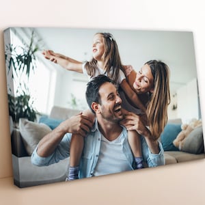 May include: A family of three, a man, a woman, and a young girl, are laughing and smiling in a living room. The girl is sitting on the man's shoulders, while the woman is holding her legs. The man is wearing a light blue shirt, the woman is wearing a white shirt, and the girl is wearing a white shirt with a pink design.