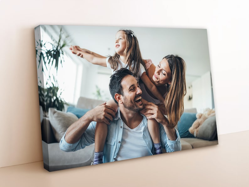 May include: A family of three, a man, a woman, and a young girl, are laughing and smiling in a living room. The girl is sitting on the man's shoulders, while the woman is holding her legs. The man is wearing a light blue shirt, the woman is wearing a white shirt, and the girl is wearing a white shirt with a pink design.