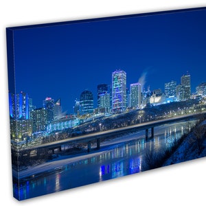 May include: A canvas print of a nighttime cityscape with illuminated skyscrapers and a bridge over a river. The buildings are lit with blue, white, and yellow lights, reflecting in the water. The sky is a deep blue.