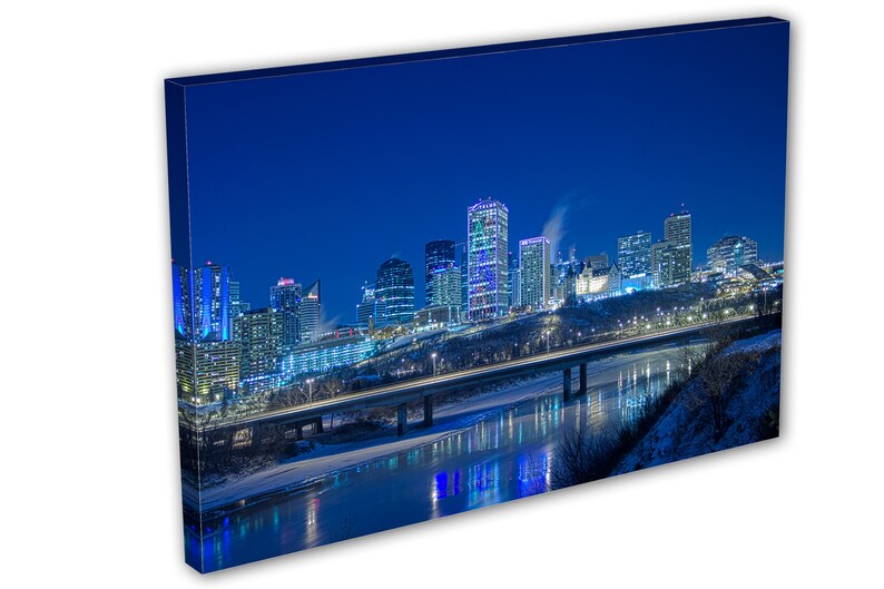 May include: A canvas print of a nighttime cityscape with illuminated skyscrapers and a bridge over a river. The buildings are lit with blue, white, and yellow lights, reflecting in the water. The sky is a deep blue.