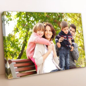 May include: A family of four, including two children, sitting on a wooden bench outdoors. The mother is holding a young girl on her back, while the father holds a young boy in his arms. The family is smiling and looking at the camera. The background is a green, leafy park.
