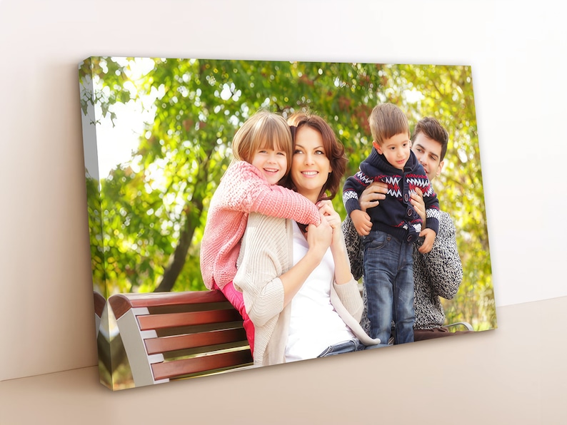 May include: A family of four, including two children, sitting on a wooden bench outdoors. The mother is holding a young girl on her back, while the father holds a young boy in his arms. The family is smiling and looking at the camera. The background is a green, leafy park.