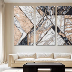 May include: A set of three abstract wall art panels with geometric designs. The artwork uses marble textures in beige, white, and dark grey, with black and gold lines. The panels are above a cream sofa and a dark wood coffee table, creating a modern interior.