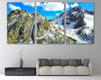 May include: A triptych canvas print of a mountain range with snow-covered peaks and patches of evergreen trees. The sky is a bright blue with fluffy white clouds. The artwork is displayed above a white sofa and a black cabinet.