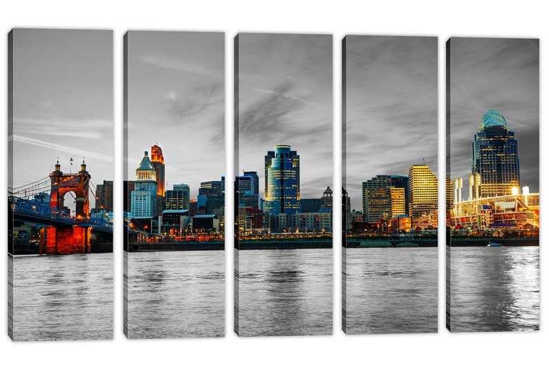 Pu&ograve; includere: Una vista panoramica dello skyline di Cincinnati al tramonto, con l'iconico ponte sospeso Roebling, il Great American Ball Park e diversi grattacieli. La citt&agrave; &egrave; illuminata da luci calde che si riflettono nel fiume Ohio.