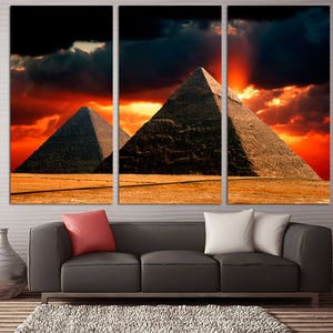 Op de afbeelding: Een driedelig canvasprint met een dramatische zonsondergang boven de Egyptische piramides. De piramides zijn gesilhouetteerd tegen een vurige oranje en rode lucht met donkere wolken.