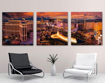 May include: A triptych of canvas prints depicting a vibrant cityscape at dusk. The artwork showcases illuminated buildings, a Ferris wheel, and a water fountain, set against a fiery orange and purple sky. Two modern chairs and a vase with a plant are in the foreground.