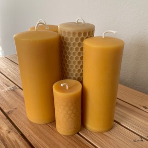 Puede incluir: Una colección de cinco velas de pilar de cera de abeja de varios tamaños. Las velas son de color amarillo cálido, algunas con un patrón de panal. Cada vela tiene una mecha blanca.
