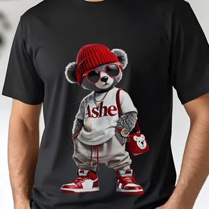 May include: Black t-shirt featuring a cartoon bear wearing a red beanie, sunglasses, and red and white sneakers. The bear is also wearing a white shirt with the word "Ashe" and carrying a red purse.