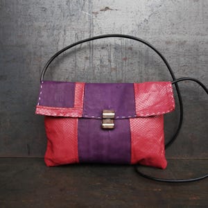 Op de afbeelding: Handgemaakte, rechthoekige crossbodytas met klepsluiting. De tas heeft een colorblock-ontwerp met rode en paarse panelen. Hij heeft een zwarte riem en een houten sluiting.