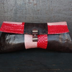 May include: A rectangular clutch with a flap closure. The clutch features a color block design with red, black, and pink sections. The clasp is a wooden rectangle with a silver accent. The bag is made of a textured material.
