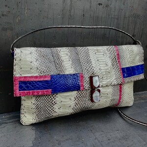 Peut inclure: Sac de type pochette avec un motif peau de serpent gris et cr&egrave;me. Le rabat pr&eacute;sente un motif color block bleu et rose. Le sac a une bandouli&egrave;re marron et une poign&eacute;e argent&eacute;e. Environ 30 cm de large.
