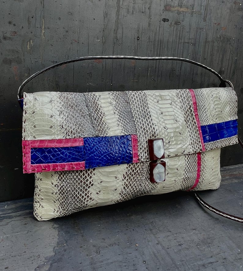 Peut inclure: Sac de type pochette avec un motif peau de serpent gris et cr&egrave;me. Le rabat pr&eacute;sente un motif color block bleu et rose. Le sac a une bandouli&egrave;re marron et une poign&eacute;e argent&eacute;e. Environ 30 cm de large.
