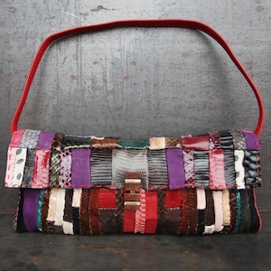 May include: A patchwork clutch bag with a red handle. The bag is made of various leather pieces in shades of red, purple, black, and white. The clasp is a rectangular gold-coloured metal.