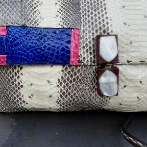 Peut inclure: Gros plan sur une pochette avec un motif peau de serpent blanc, gris et marron. Le sac &agrave; main pr&eacute;sente un accent bleu et rose, avec deux fermoirs rouge fonc&eacute; avec des inserts en coquillage blanc. Une sangle marron est visible.