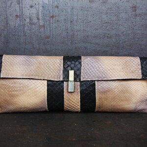 Könnte beinhalten: Rechteckige Clutch mit Schlangenmuster in Beige und Schwarz. Die Clutch hat einen Klappenverschluss mit silberfarbener Schnalle. Die Enden der Clutch sind schwarz, die Mitte hat einen schwarzen Streifen.