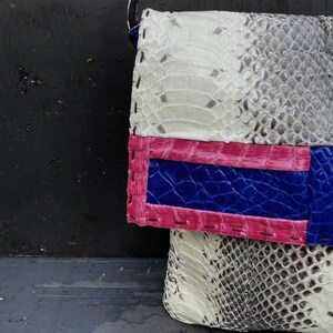 Peut inclure: Sac &agrave; main patchwork fait &agrave; la main avec une bandouli&egrave;re noire. Le sac pr&eacute;sente un motif peau de serpent blanc, gris et marron. Un panneau rose vif et bleu est cousu sur le devant, ajoutant une touche de couleur au design.