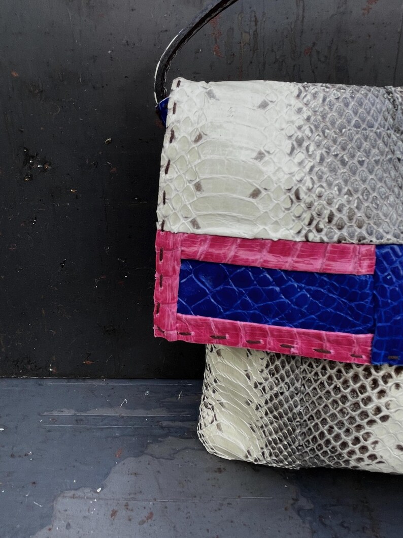 Peut inclure: Sac &agrave; main patchwork fait &agrave; la main avec une bandouli&egrave;re noire. Le sac pr&eacute;sente un motif peau de serpent blanc, gris et marron. Un panneau rose vif et bleu est cousu sur le devant, ajoutant une touche de couleur au design.