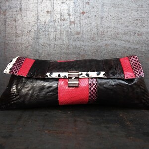 May include: A black clutch with red and white accents. The clutch features a textured, reptile-like pattern. The design includes a red stripe across the center and a white and black patterned flap. The clasp is silver and brown.