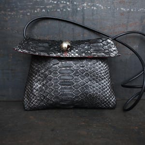 Pode incluir: Uma pequena bolsa de couro preta com alça transversal, com uma fivela prateada e um padrão de pele de cobra.