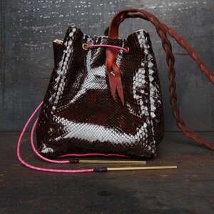 Peut inclure: Un sac seau marron et blanc à motif peau de serpent avec une bandoulière en cuir tressé marron. Le sac a une fermeture à cordon rose et deux tiges métalliques dorées. Le fond est une surface sombre et texturée.