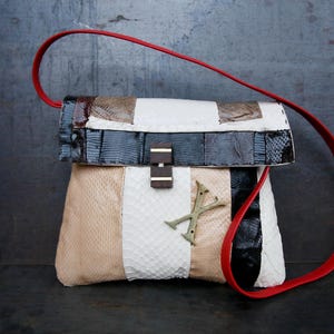 May include: A patchwork handbag with a red strap. The bag features panels of beige, white, black, and patterned leather. A gold-coloured metal emblem and a wooden clasp adorn the front.