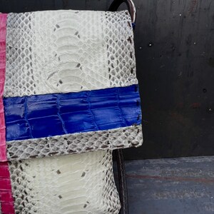 Peut inclure: Un sac &agrave; main de type pochette fait &agrave; la main avec un design patchwork. Le sac combine du cuir textur&eacute; blanc, rose et bleu. La section blanche pr&eacute;sente un motif peau de serpent. Le sac a une sangle noire.