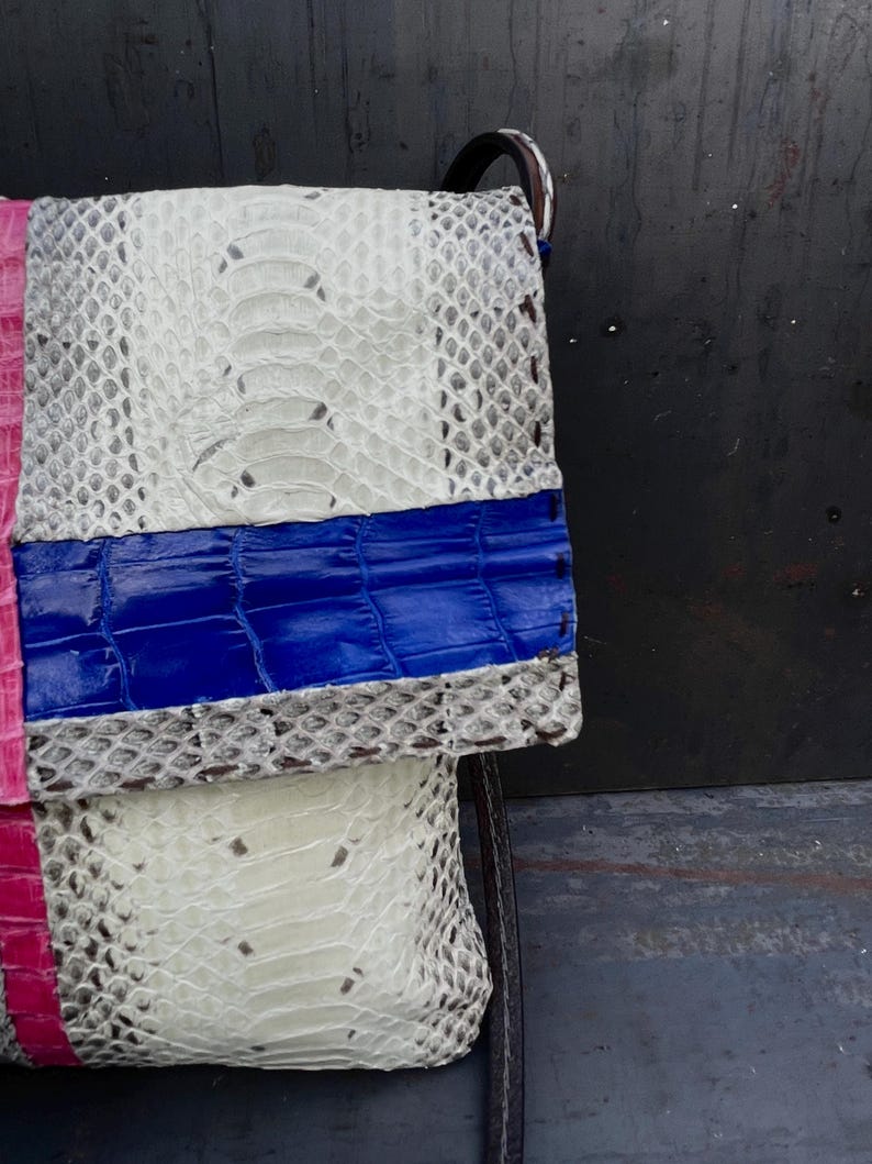 Peut inclure: Un sac &agrave; main de type pochette fait &agrave; la main avec un design patchwork. Le sac combine du cuir textur&eacute; blanc, rose et bleu. La section blanche pr&eacute;sente un motif peau de serpent. Le sac a une sangle noire.