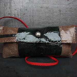 May include: A rectangular clutch bag with a snakeskin pattern in shades of brown, black, and white. The bag has a silver button closure and a red braided strap. The bag is designed to be carried over the shoulder.