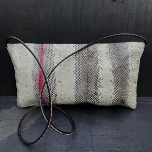 Peut inclure: Une pochette rectangulaire avec un motif peau de serpent en blanc, gris et noir, accentu&eacute;e d'une bande rose vif. La pochette est dot&eacute;e d'une longue bandouli&egrave;re en cuir noir pour un port&eacute; &eacute;paule ou travers.