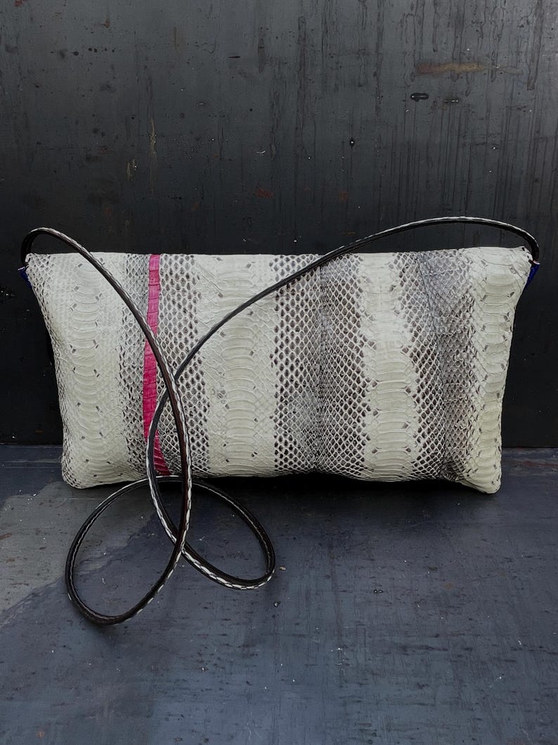 Peut inclure: Une pochette rectangulaire avec un motif peau de serpent en blanc, gris et noir, accentu&eacute;e d'une bande rose vif. La pochette est dot&eacute;e d'une longue bandouli&egrave;re en cuir noir pour un port&eacute; &eacute;paule ou travers.
