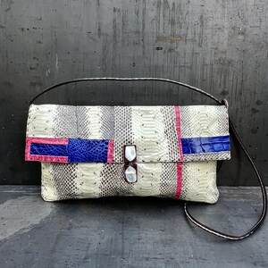 Peut inclure: Une pochette rectangulaire avec un motif peau de serpent blanc, gris et noir. Le rabat pr&eacute;sente des accents roses et bleus. Le sac a une sangle noire et une fermeture &agrave; double fermoir.