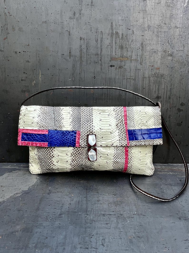 Peut inclure: Une pochette rectangulaire avec un motif peau de serpent blanc, gris et noir. Le rabat pr&eacute;sente des accents roses et bleus. Le sac a une sangle noire et une fermeture &agrave; double fermoir.