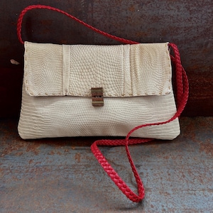 Op de afbeelding: Een witte leren clutch met een rode gevlochten band. De tas heeft een klep sluiting met een metalen sluiting.