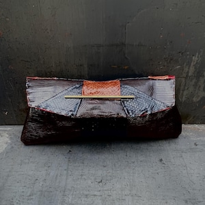 May include: A dark brown clutch with a reptile-like texture. The clutch has a rectangular shape with a fold-over flap. A gold-colored bar accents the front. The top section is a lighter brown color. The edges are trimmed with red.