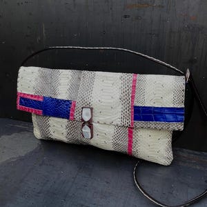 Peut inclure: Pochette en peau de serpent blanche, grise et noire, avec une bandouli&egrave;re noire. Le rabat est orn&eacute; d'accents bleus et roses, et le fermoir est en nacre.