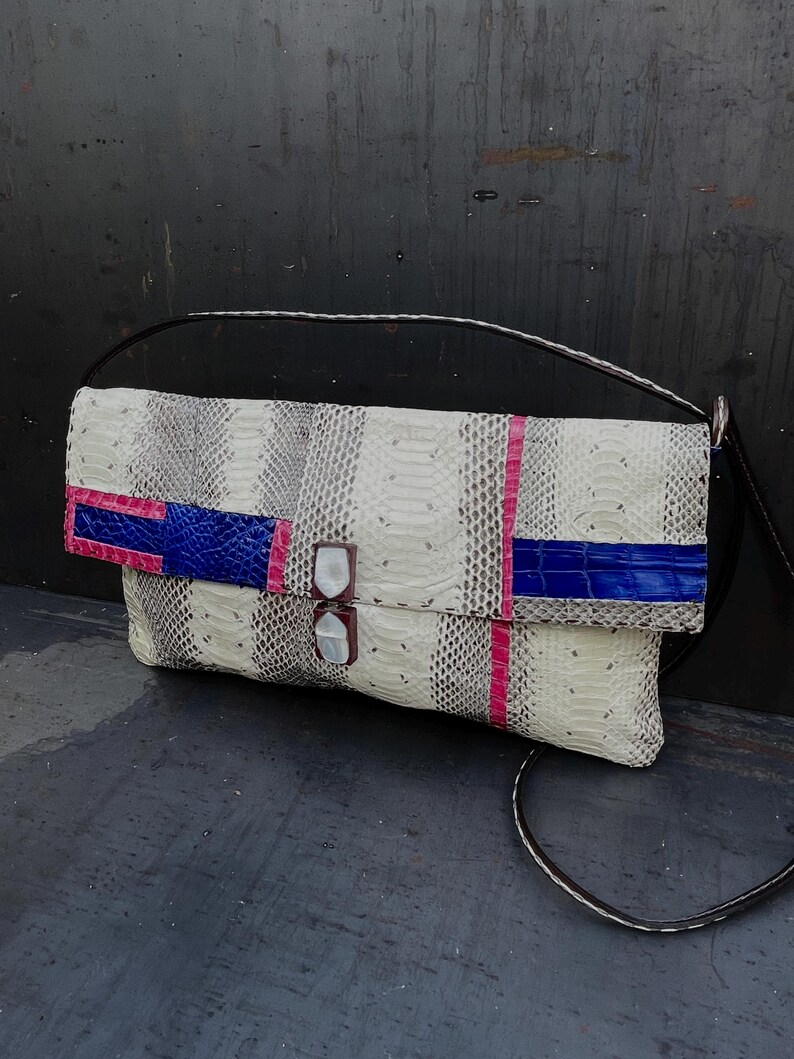 Peut inclure: Pochette en peau de serpent blanche, grise et noire, avec une bandouli&egrave;re noire. Le rabat est orn&eacute; d'accents bleus et roses, et le fermoir est en nacre.