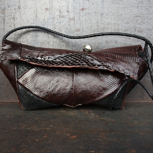 Könnte beinhalten: Eine braune Ledertasche mit einem schwarzen Riemen und einem silbernen Verschluss. Die Tasche hat ein strukturiertes, schlangenhautähnliches Muster.