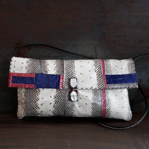 Peut inclure: Pochette rectangulaire avec un motif peau de serpent gris et blanc. Le rabat présente des accents rouges et bleus. Le sac a une bandoulière noire et une fermeture à fermoir.
